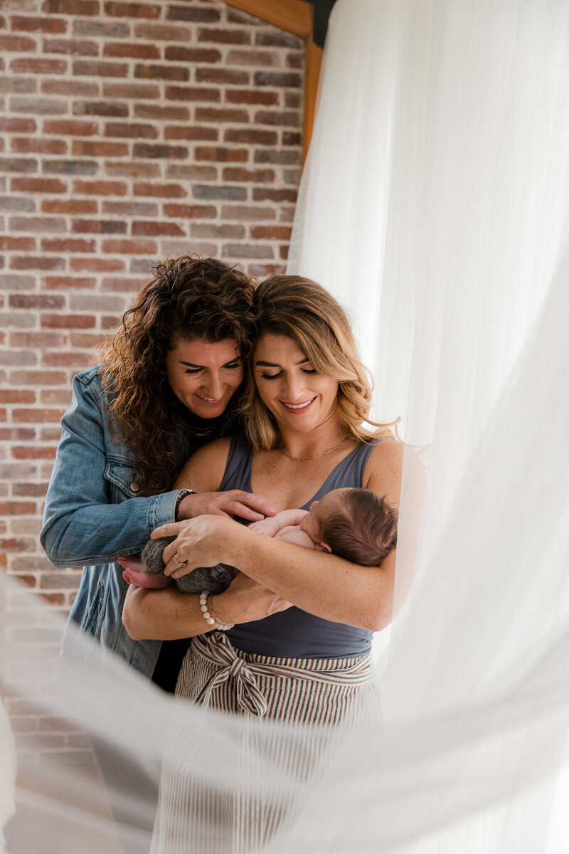 Lesbian couple holding newborn baby boy by window with white curtains - inclusive family newborn photography by Glean and Co Boise studio