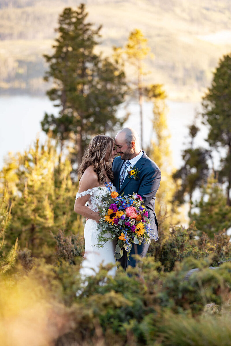 A newlywed couple standing forehead to forehead in a wooded area.