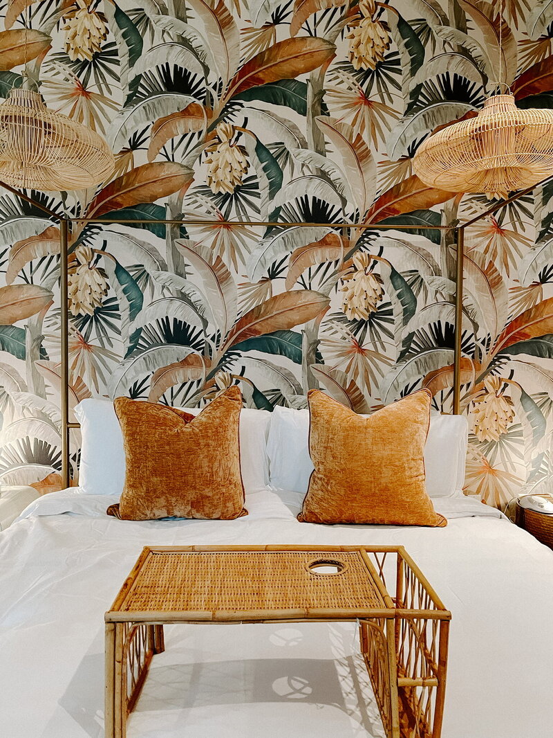 white bed with orange accent pillows against wallpaper of palm leaves