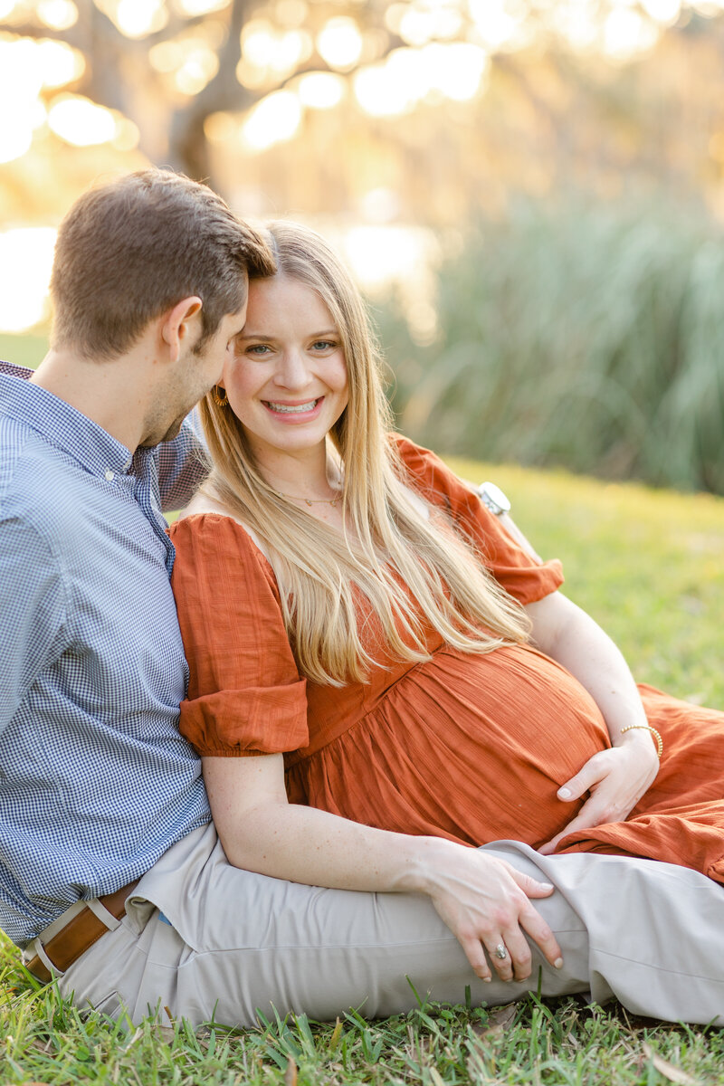 Pregnant mother smiles at camera in orange dress while lounging in the grass with her husband in Tampa, Florida