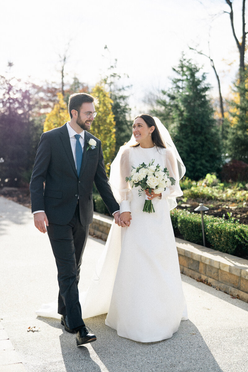 Bride in a classic long-sleeved gown and groom in a dark suit walking hand in hand outdoors, smiling at each other on a sunny fall day surrounded by greenery.