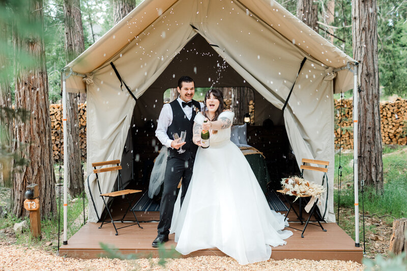 A bride pops champagne while standing with her groom in front of a canvas tent cabin on their Yosemite elopement day. 