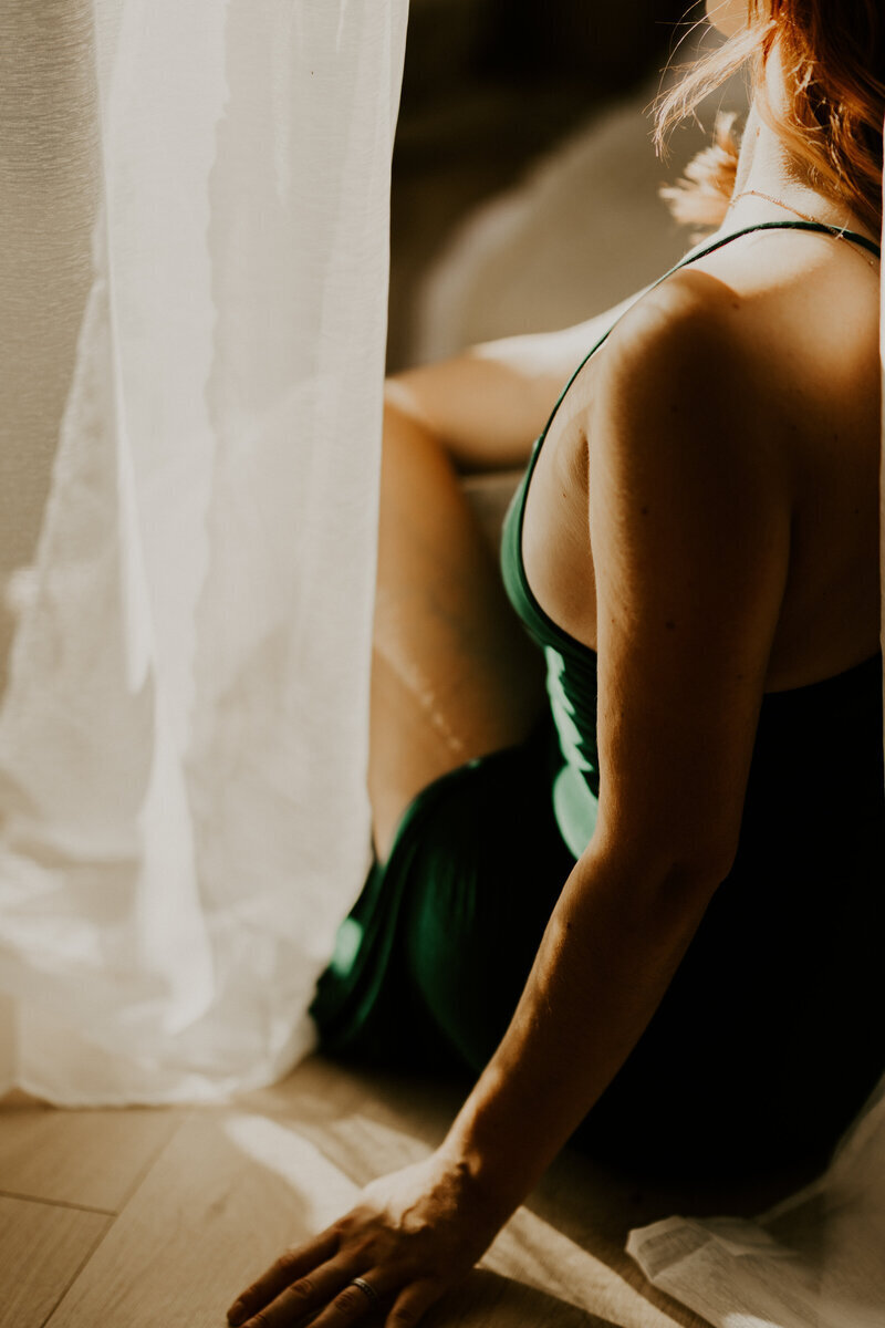 Femme rousse assise près d'un voilage blanc, robe verte, lumière douce sur son épaule créant une atmosphère intime, prise au studio de Laura, photographe en vendée.