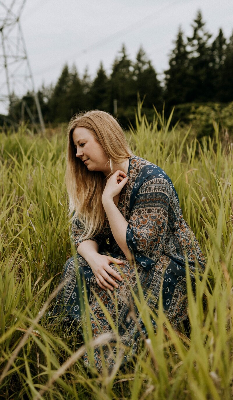 Jaymie Sutherland, Spiritual Healer, Activator, & Expander, standing with a bridge in the background, looking over to the left with a hands clasped in front of her & a soft smile on her face.