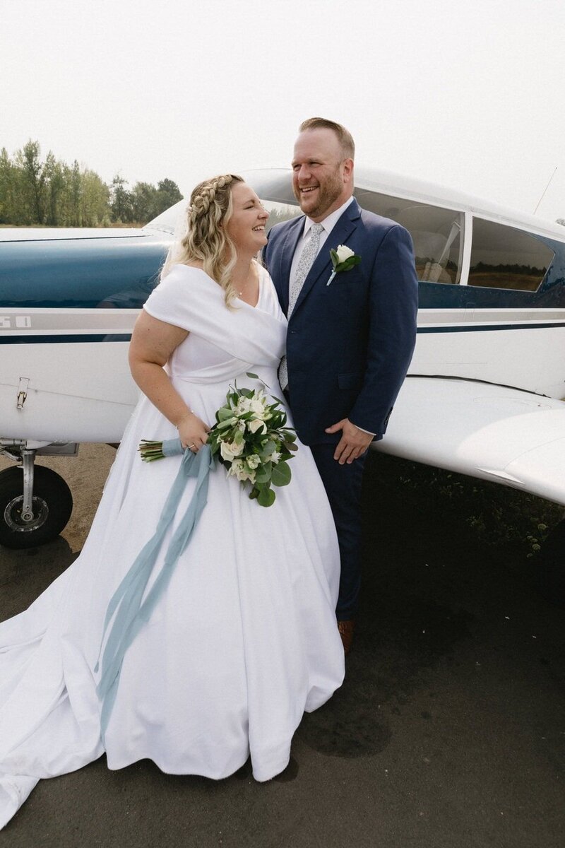 Newlyweds stand in front of the groom's hobby plane and look towards each other.