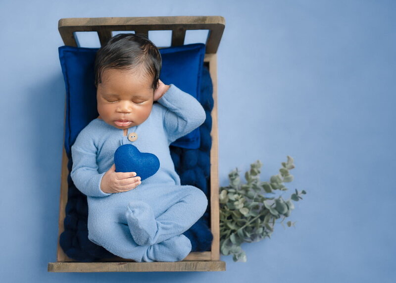 Baby boy in blue sleeper holding heart plush, posed in wooden bed during newborn mini session in Birmingham studio
