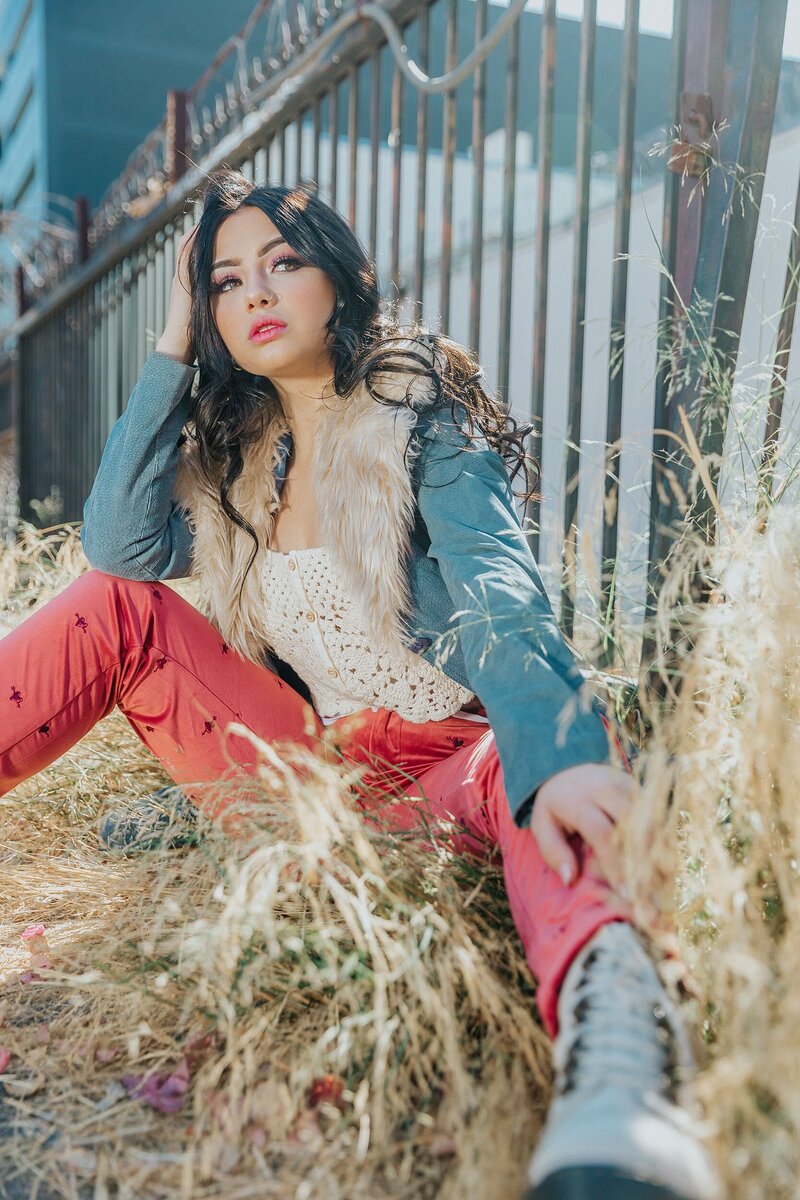 Bold teen model in a fur-trimmed jacket and coral pink pants, sitting in tall grass near a metal fence during a styled mentorship session with Tara Rochelle.