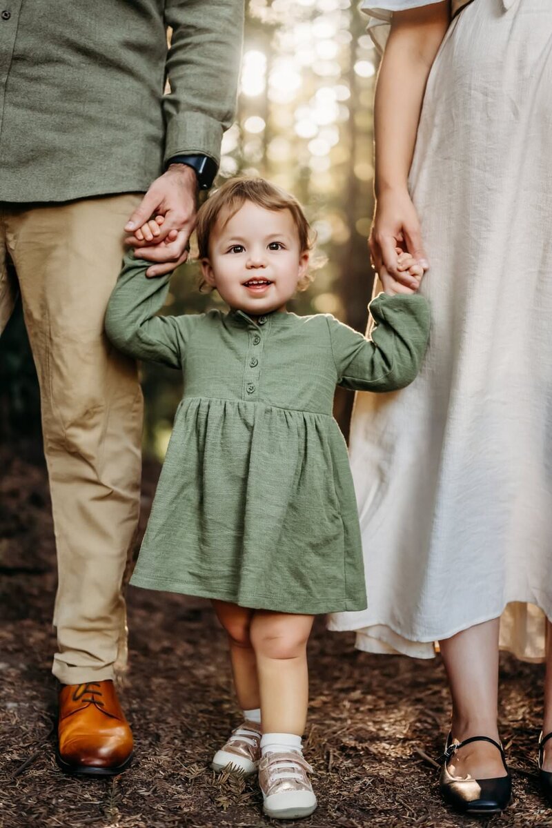 toddler holds parents hands as sun filters through redwood forest behind them
