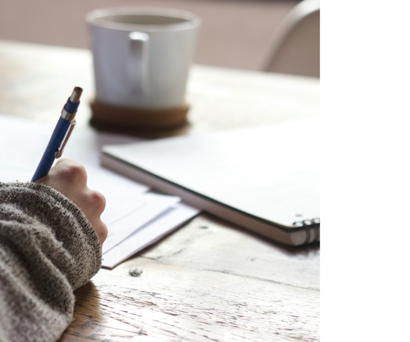 image of a hand holding pen writing on piece of paper with coffee cup on desk