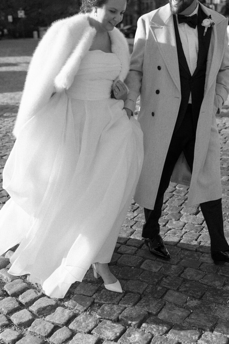 Black and White image of bride and groom crossing cobblestone street. Captured by Caitlin Wilcox Photography, wedding photographer in Richmond VA