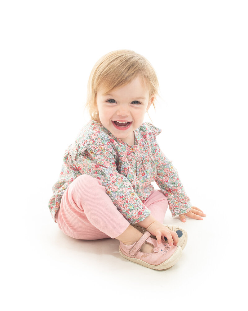 A toddler sits on the floor in a bright studio at Dandelion Studio in Moira, Northern Ireland. Dressed in pink trousers and a floral top, the toddler smiles with a joyful expression, showing curiosity and energy.