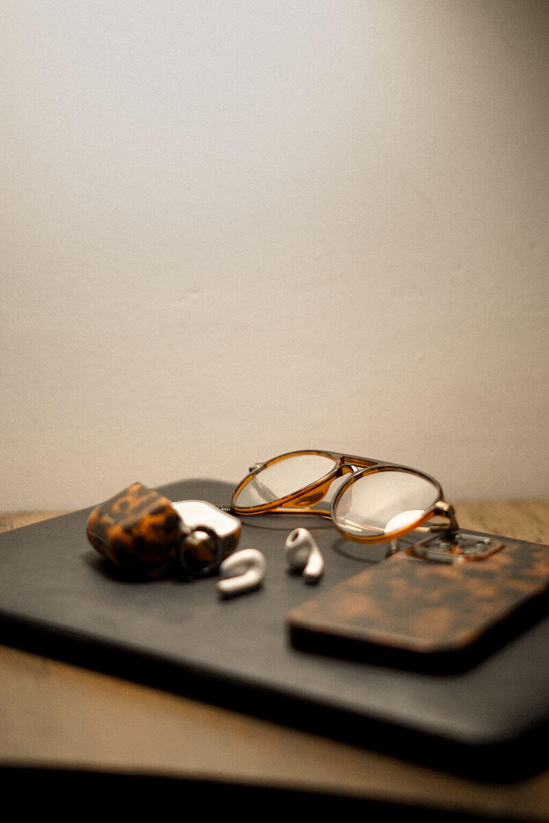 A pair of eyeglasses, wireless earbuds, a patterned case, and a matching phone case by Sweet Pea Ink Creative rest on a dark surface against a plain background.