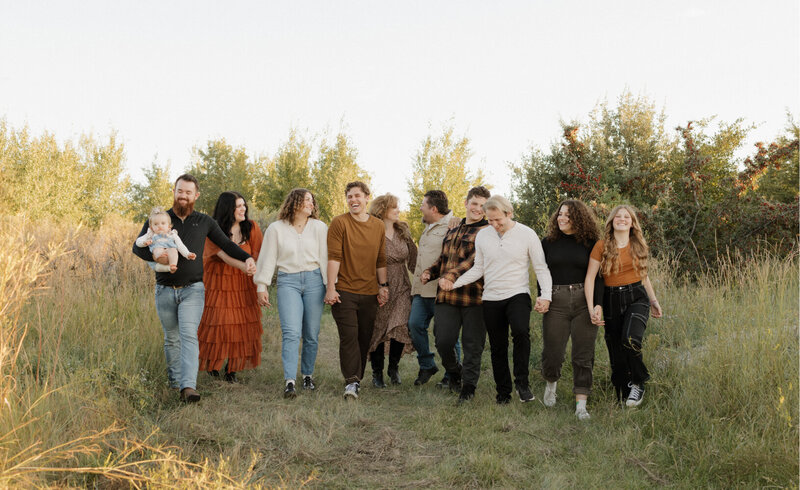 A large family consisting of 10 people and a baby all holding hands, walking towards the camera, and smiling at each other outside.