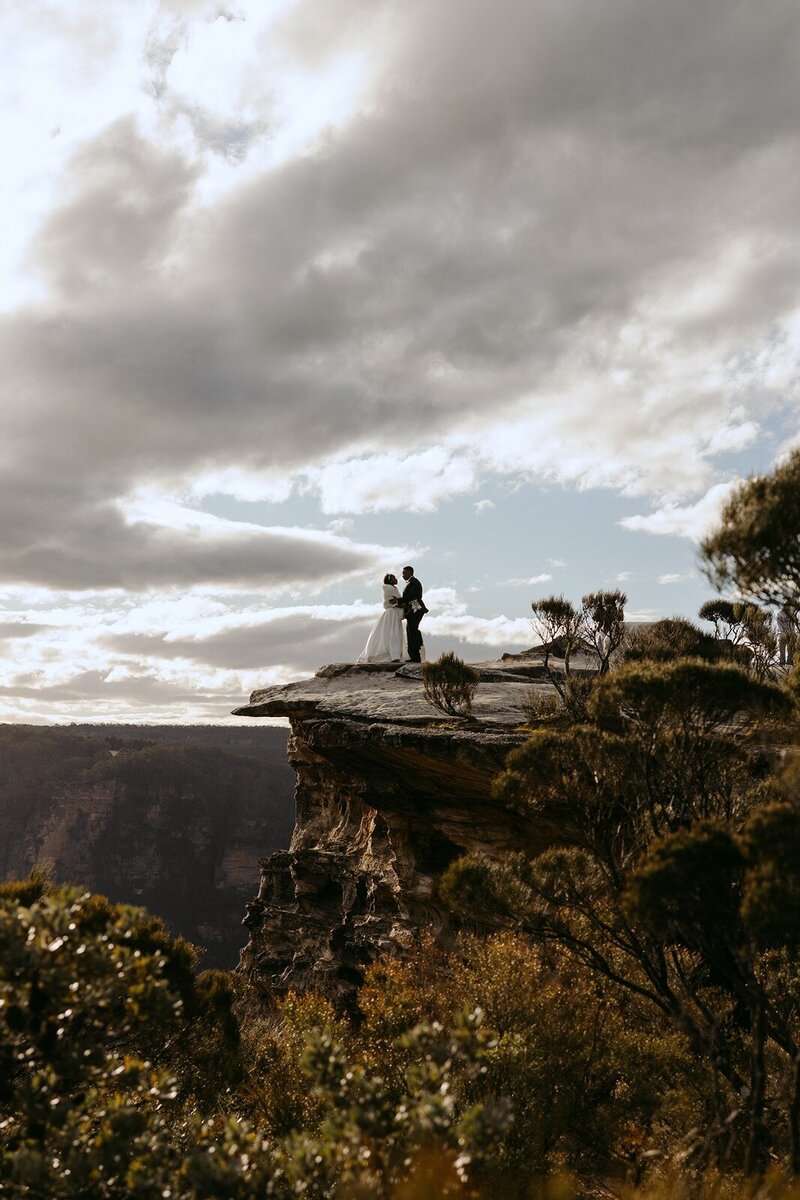 A wide shot of a bride and groom on a cliff in the blue mountains