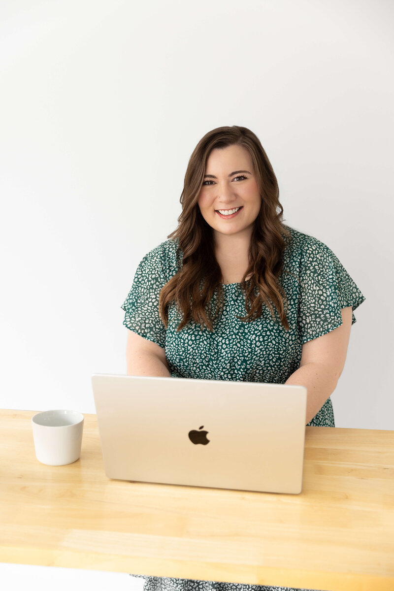 Brand and web designer smiling at her desk with a laptop and coffee mug, working in a bright minimalist office.