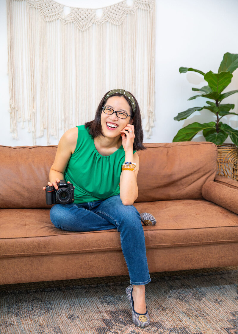 An Asian woman sitting on a brown couch holding a Nikon D750 camera.