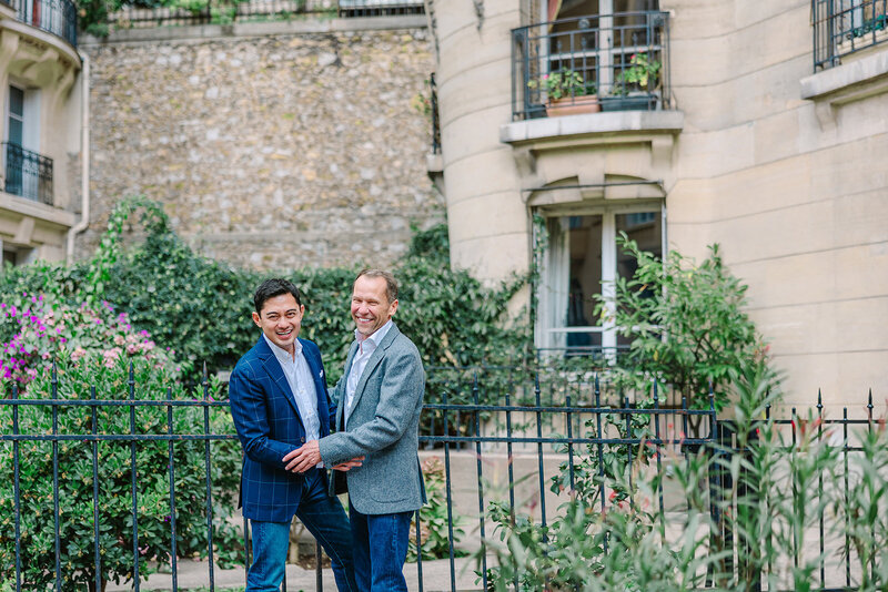 Gay couple holding hands and laughing at the camera near the Eiffel Tower at Bir-Hakeim Bridge during a romantic Paris couples photoshoot.