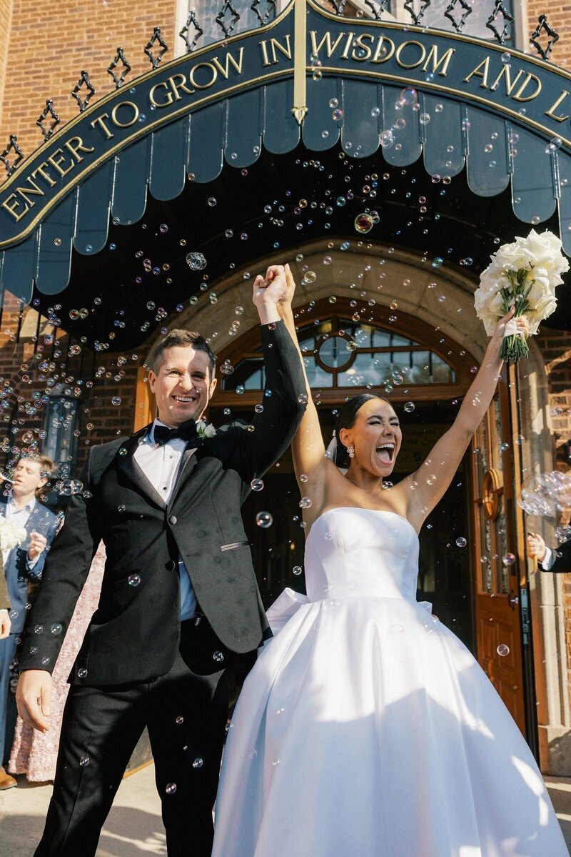 bride and groom on wedding day excitedly raising arms while exiting holding bouquet and bubbles blown into frame shot by minnesota photographer tom thornton photography