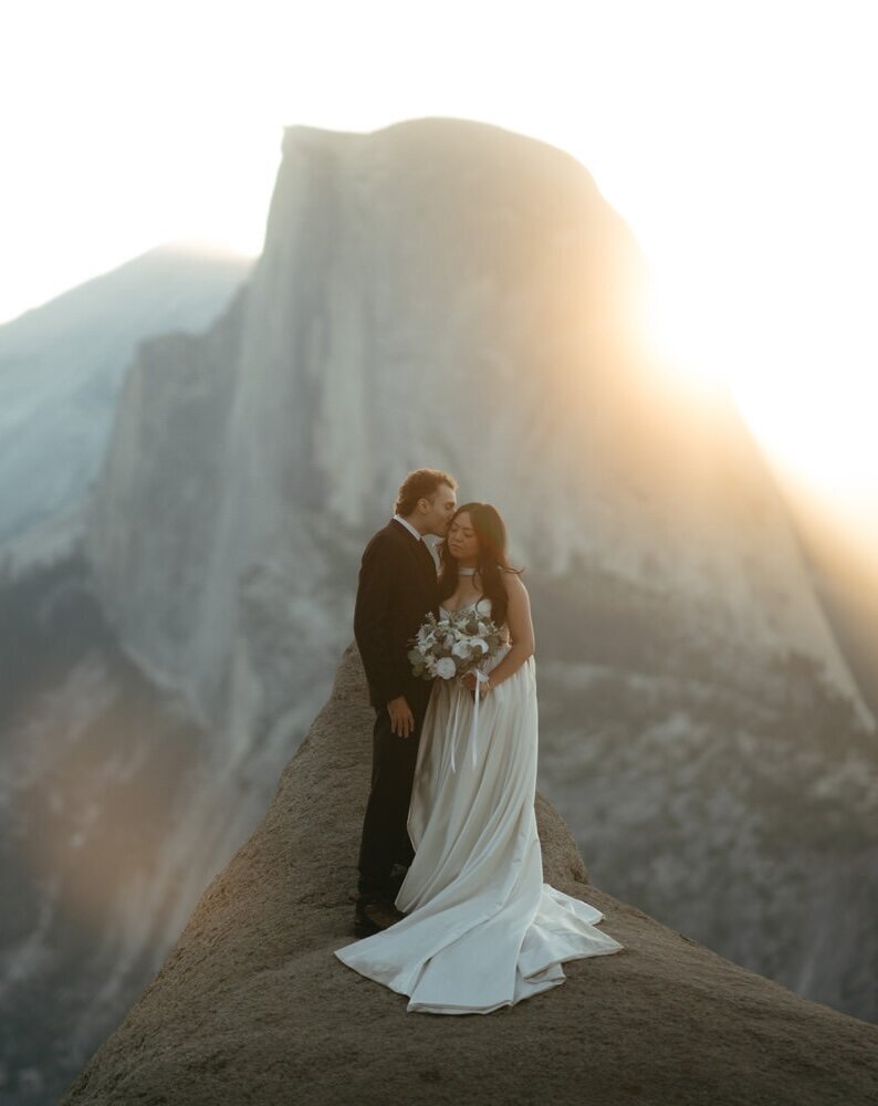 Groom kisses bride on the cheek with a large mountain in the background of their hiking wedding trail