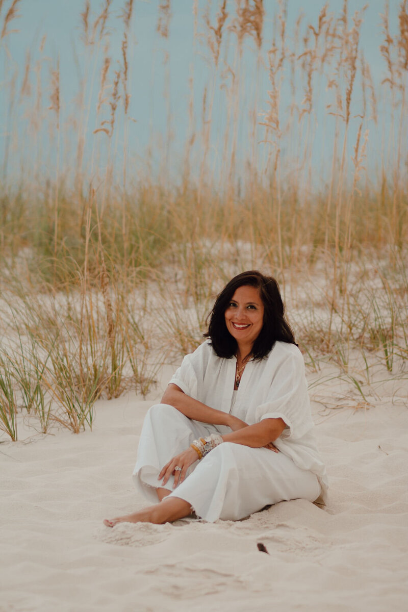 Smiling woman in white pants and white shirt with lots of bracelets on her art with shoulder length dark brown hair sitting in the sand with dune grass behind her