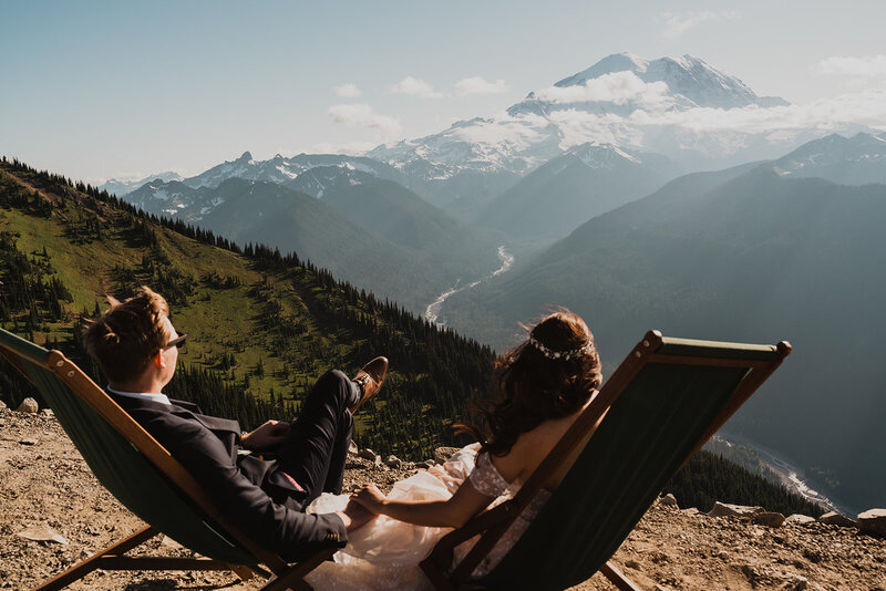 a couple, in their wedding attire sit in camp chairs gazing out at the mountains ahead of them. 