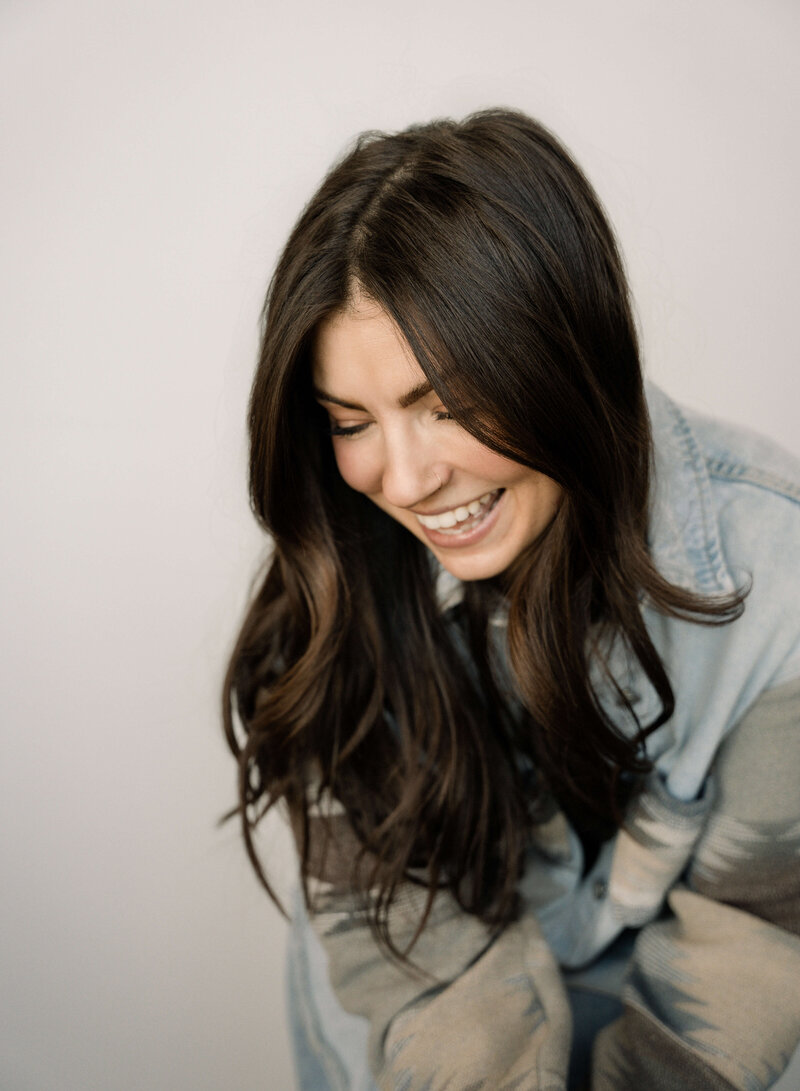 A woman laughs as she looks down to her right with long brown hair and a denim jacket
A woman laughs as she looks down to her right with long brown hair and a denim jacket