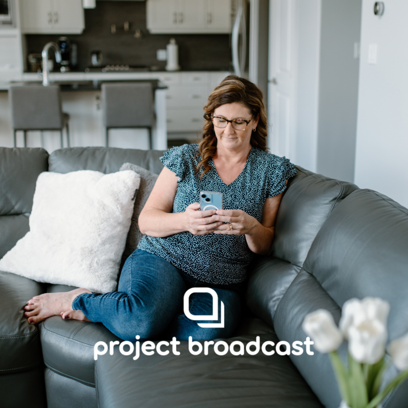 Michelle, founder of The Systems Café, sitting comfortably on a gray couch with her smartphone, showcasing the use of Project Broadcast for streamlined and personalized text marketing, with the Project Broadcast logo overlayed