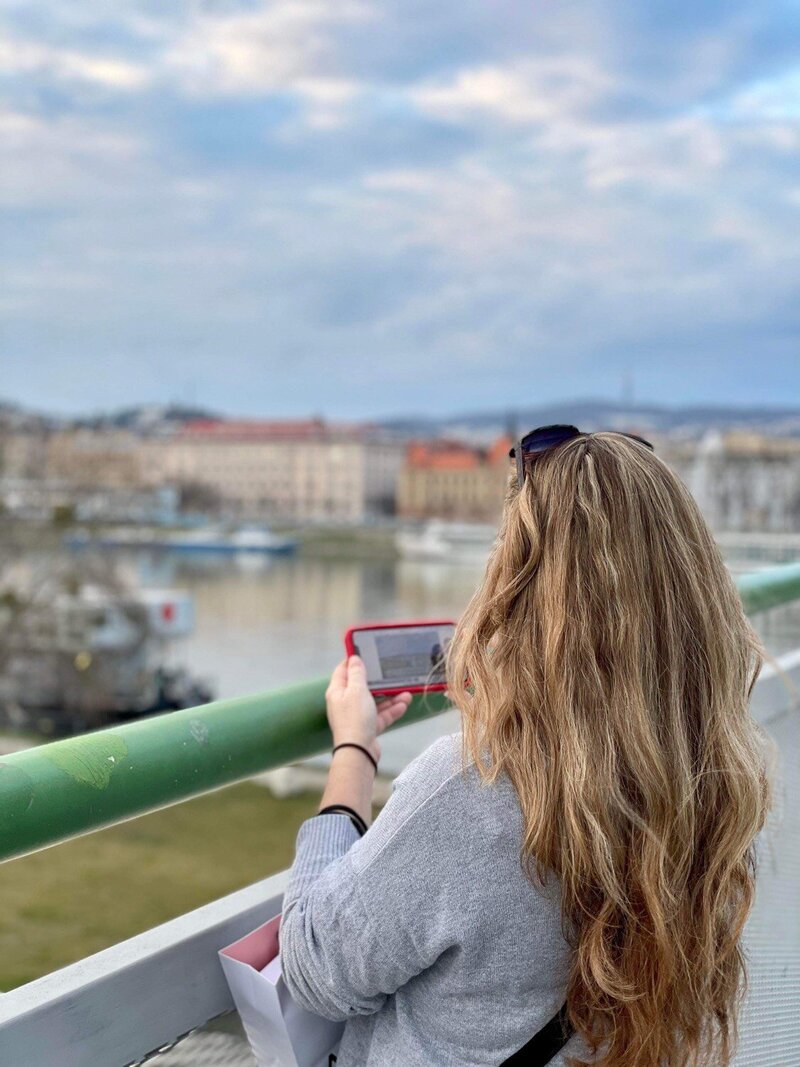 Terra from Adventures by Terra standing on a bridge above the Danube river taking photos on her phone in Bratislava, Slovakia