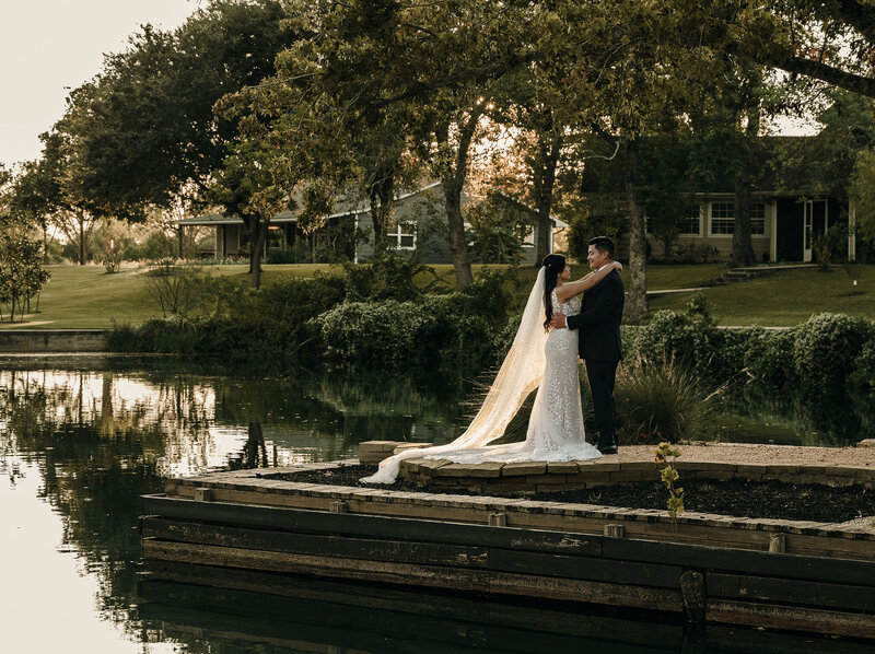 A bride and groom stand on a dock by a serene lake, embraced and gazing at each other. The bride's veil flows behind. Trees and a house are in the background.