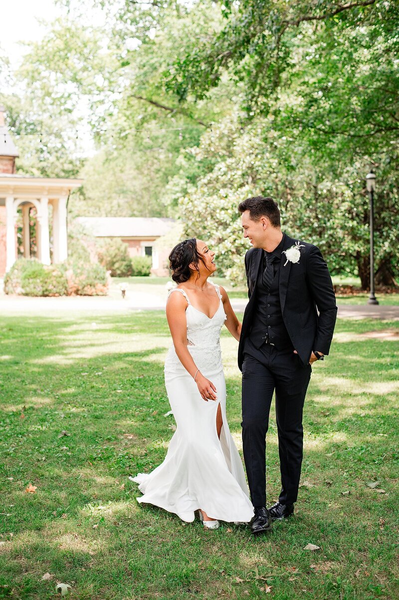 Bride wearing a fitted wedding gown with a flaired skirt and thigh high slit laughs up at the groom who is holding her hand. The groom is wearing a black suit with a black shirt and a white rose boutonniere. They are walking the grounds at Oaklands Mansion.