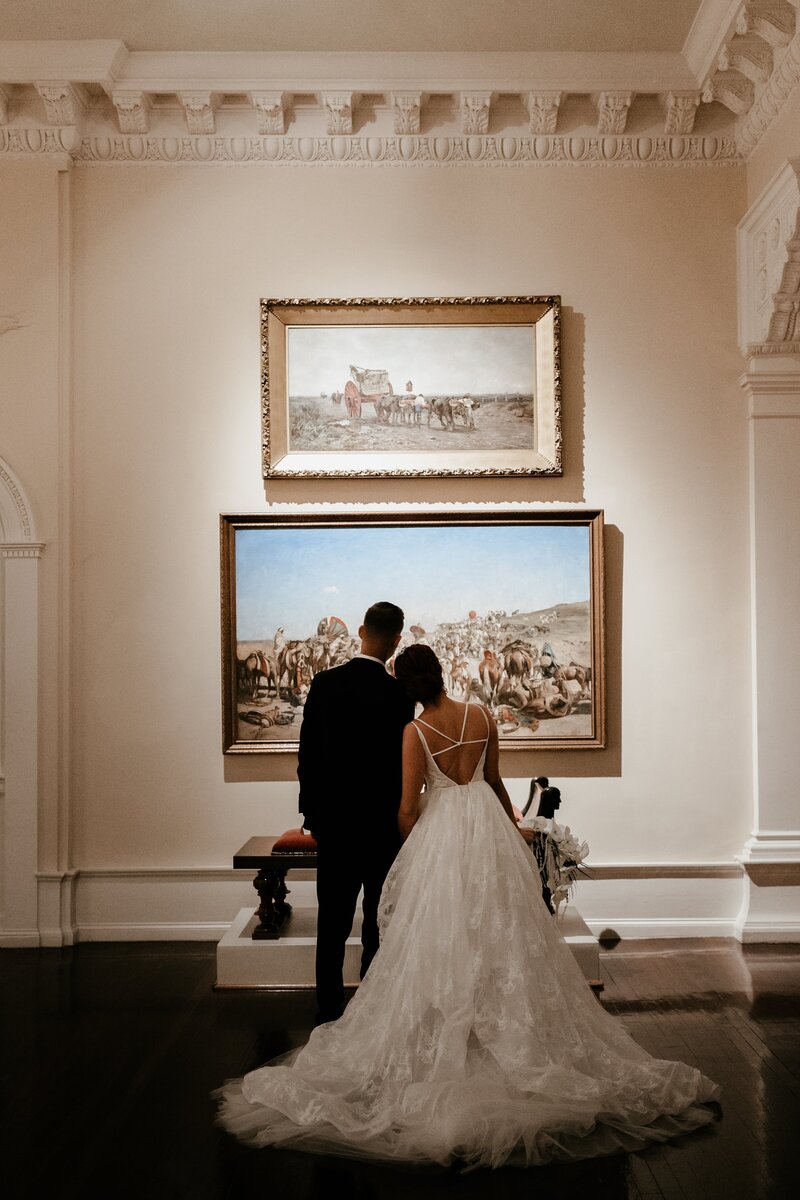 A bride and groom with their backs to the camera looking at a painting of cattle