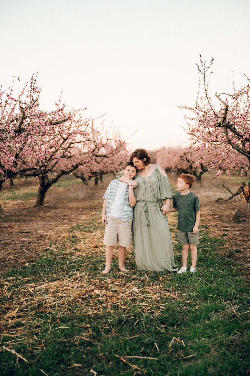 Melissa, a Family Photographer in Macon GA stands with her two children in a blooming orchard, surrounded by pink spring blossoms.