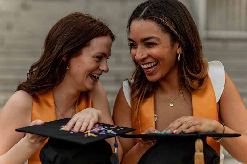 2 college grads hold confetti on their caps while they giggle together
