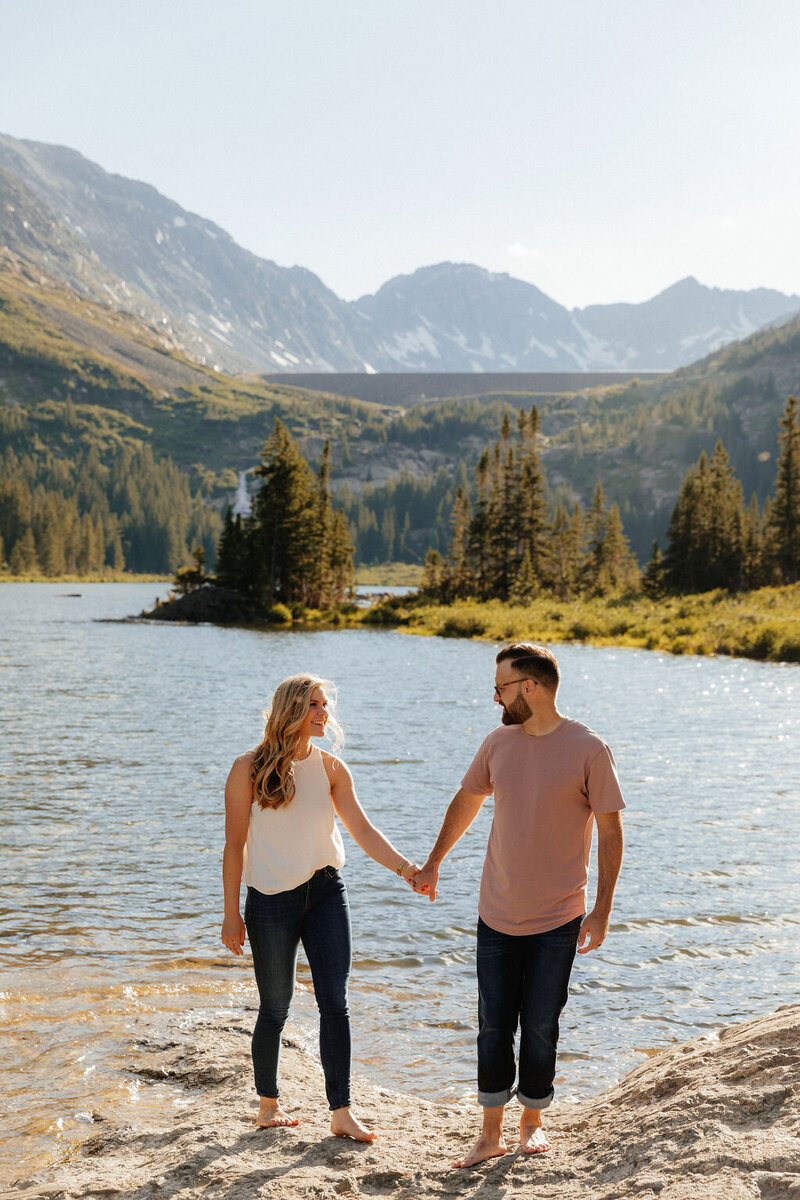 Couple holding hands in the mountains of Breckenridge walking barefoot through an alpine lake for their summer engagement session