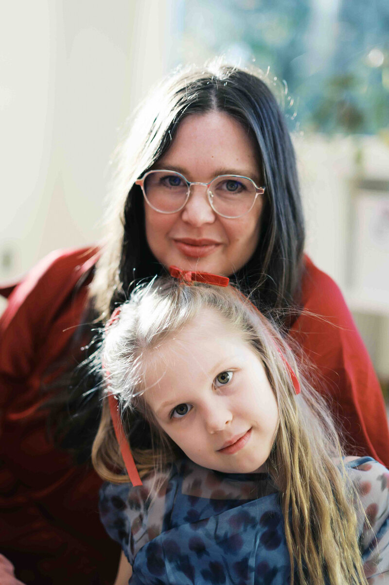woman wearing red dress with dark brown hair and trendy glasses rests chin on head of young girl wearing blue dress with red ribbons in blonde hair by Portland Family Photographer Emilie Phillipson Photography. 