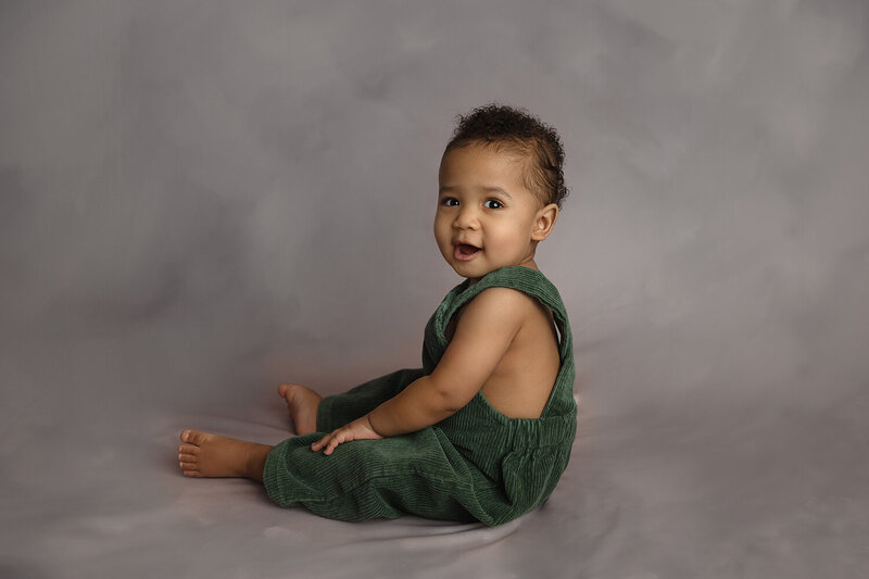 A young toddler sits on the floor of a studio in green overalls with a big smile
