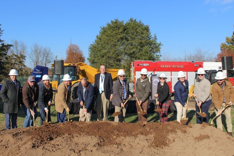 A group of people wearing hard hats and holding shovels participate in a groundbreaking ceremony. They are lined up in front of construction equipment and a fire rescue vehicle, digging into a mound of dirt.