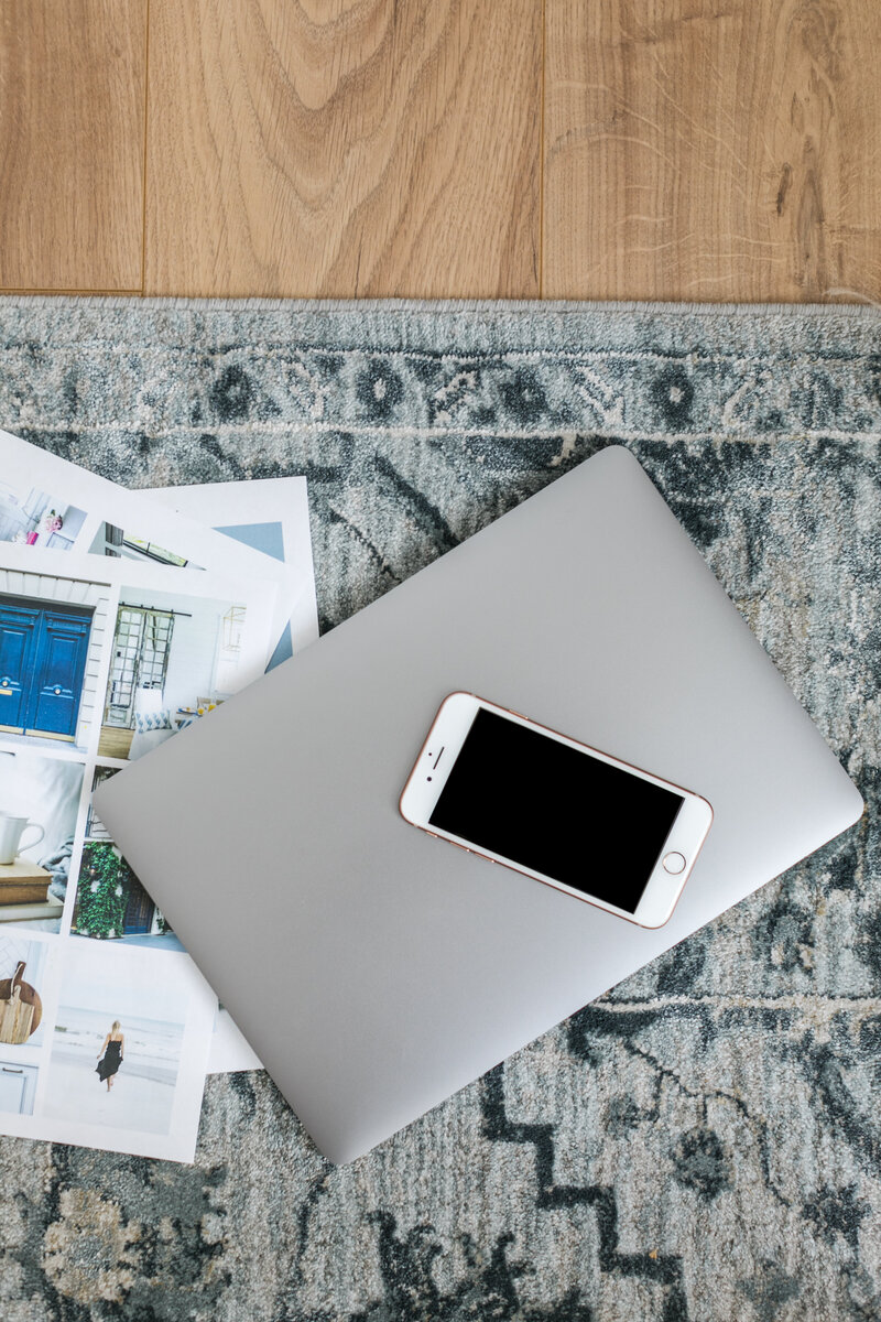Overhead photo of a closed silver laptop with a white iPhone on top, resting on a blue rug next to design inspiration printouts. Pairs with copy about the chaos of solo business ownership — from missed appointments to late-night workflow tweaks — and emphasizes the need for simple, tailored systems for women entrepreneurs.