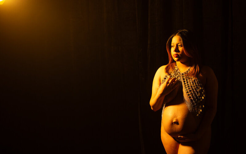 contemplative maternity portrait in studio on a black backdrop with golden lighting to give her a glow, hispanic mom to be andreina is wearing a sparkling metal top with her belly as the focus facing forward. charlotte nc