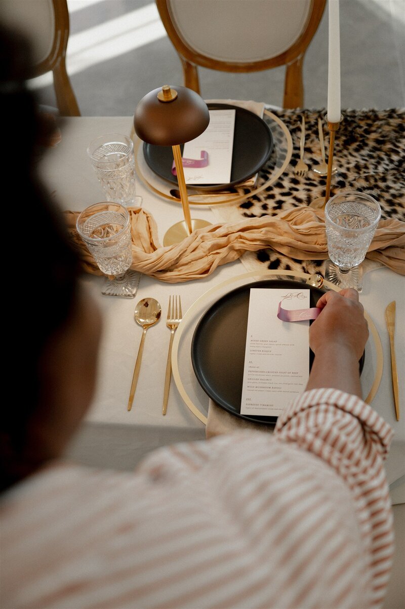 Grey Cove Designs dinner menu styled on a modern black plate with gold flatware, knotted silk runner, and crystal glassware, as a guest lifts a ribbon-wrapped menu at an intimate wedding table.