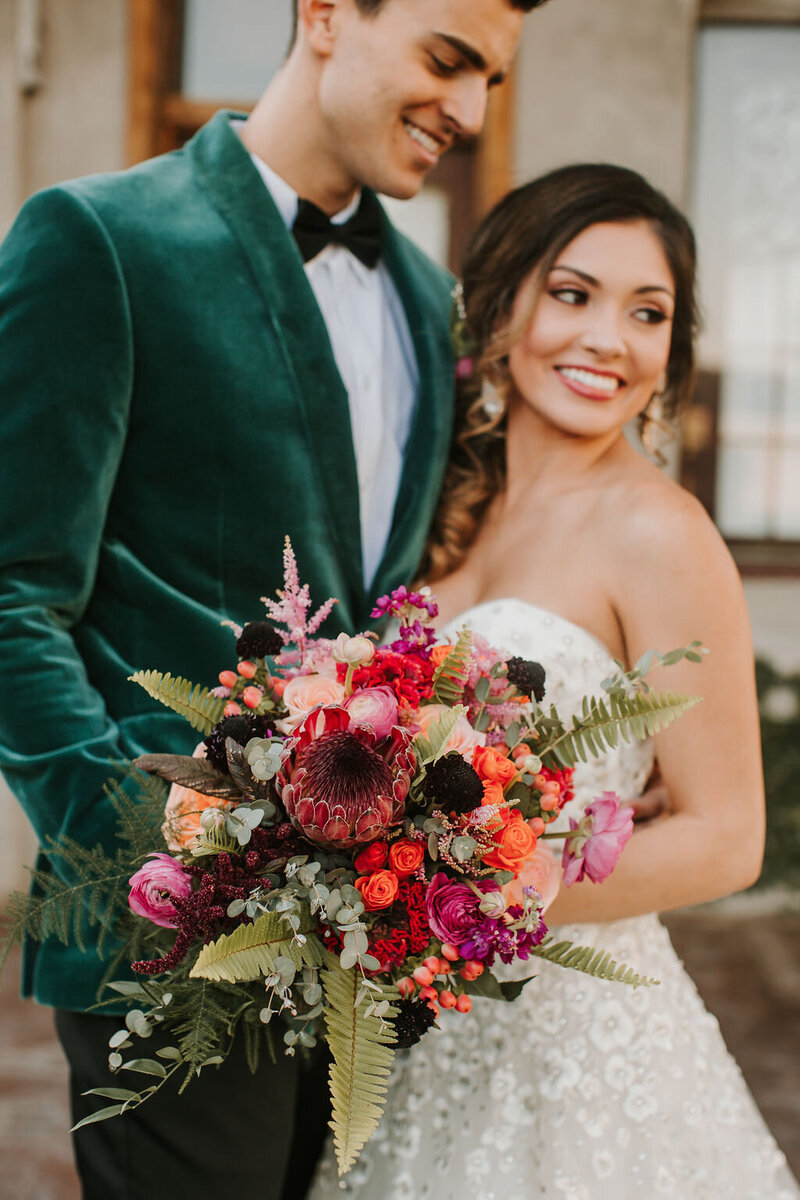 Bride and groom with their arms interlocked smiling at each other in front of a brick wall