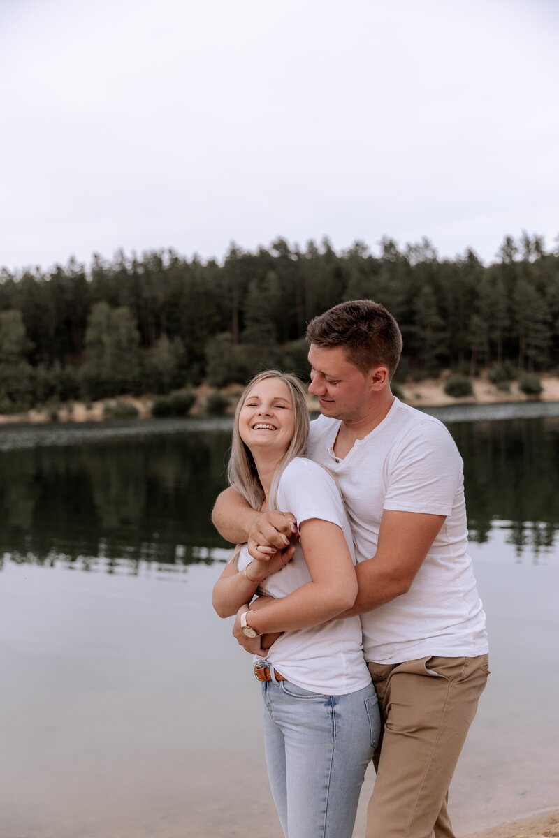 Mann und Frau stehen an einem See er umarmt sie von hinten
