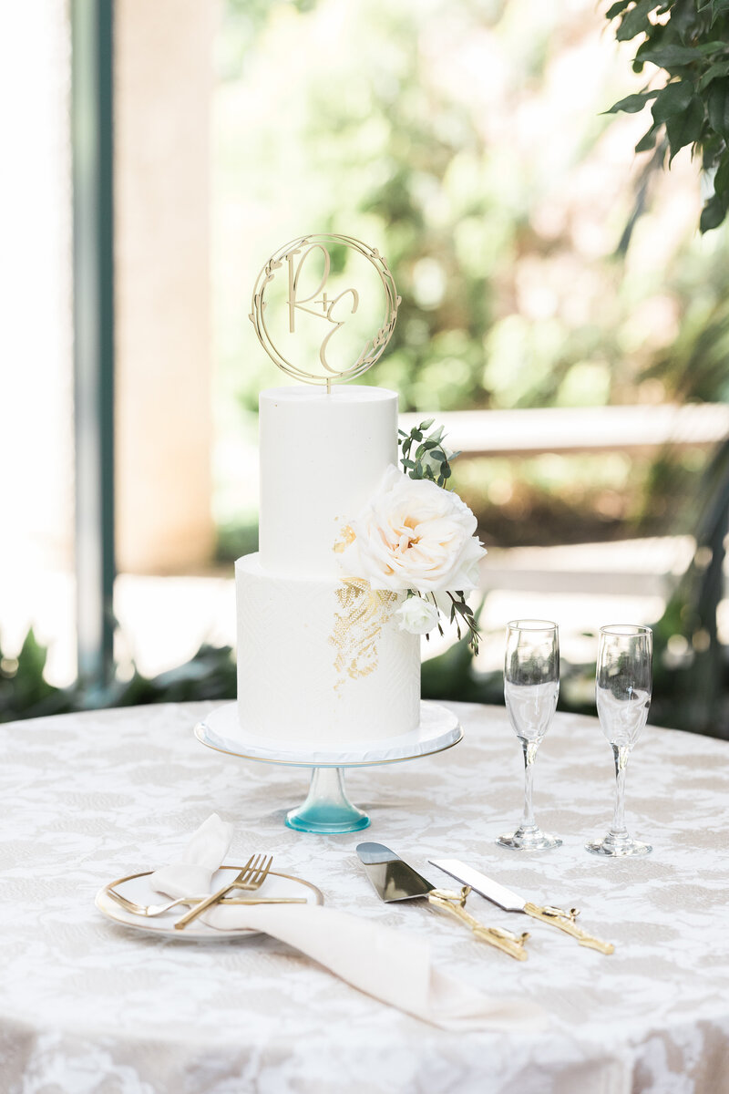 textured dusty blue and gold flecked floral linen with a 2-tier white buttercream cake with a gold wood cake topper "R+E" sitting on a glass clear and blue cake stand with two champagne flutes, gold cake cutlery, and a white and gold rimmed cake plate with gold forks 