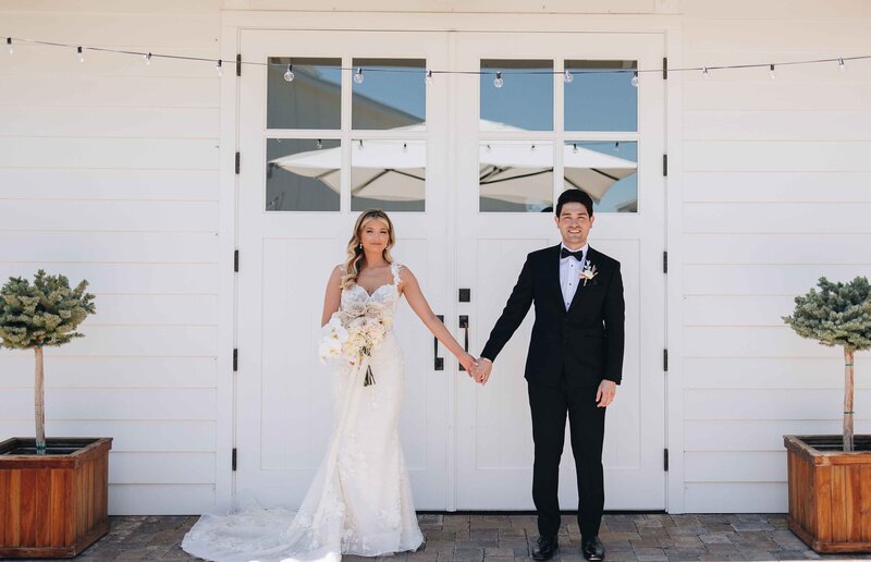 Couple Holding Hands in Front of Lux White Barn - Mikayla & Mario | Harmony Meadows Wedding - Lake Chelan Wedding