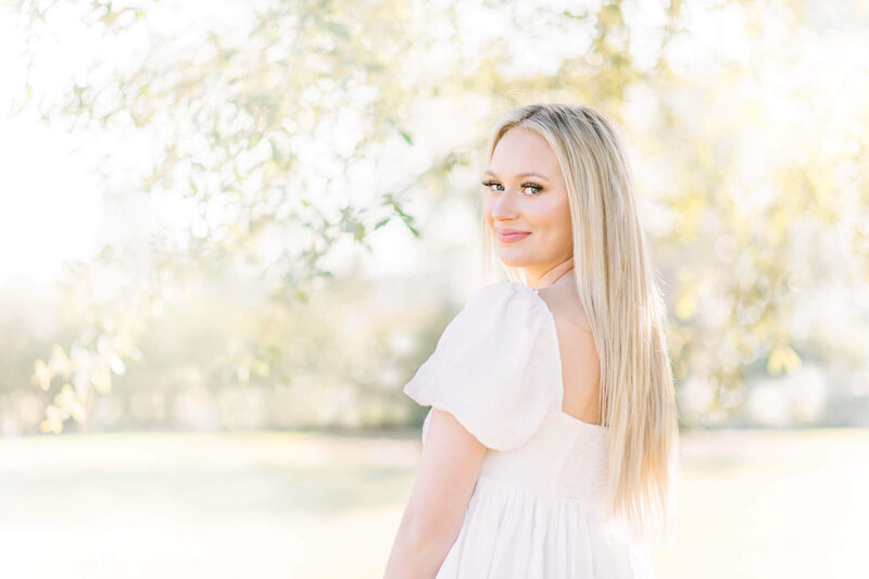 A blonde Albany High School senior wearing a white dress looks back over her left shoulder in a field under a tree on a sunny day; taken by Morgan Leigh Photography