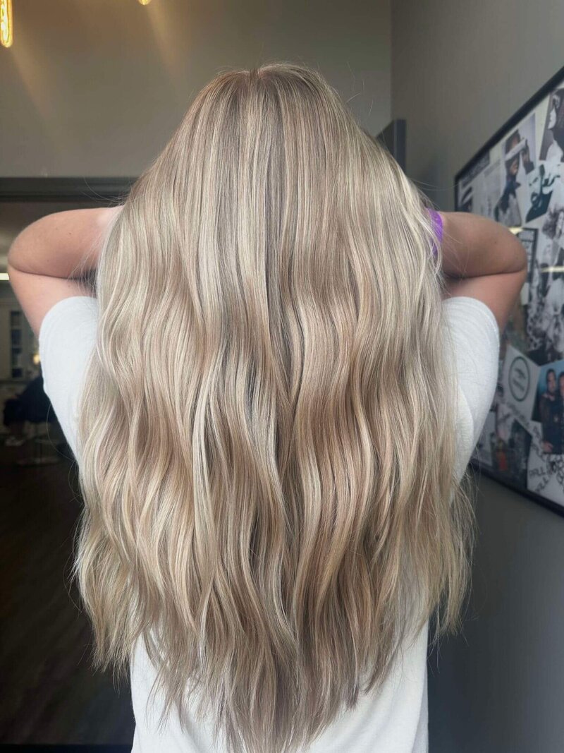 A woman with blonde hair, slightly curled, facing away from the camera, showcasing the elegant styling done by 212 Salon, Spa, & Barbershop..