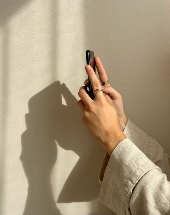 A young woman with auburn hair and a playful smile holds a professional camera with a large lens. She wears a cropped white long-sleeve shirt and beige high-waisted trousers. Her left hand is raised toward the camera capturing her, partially obscuring her face. A curtain rail with a hanging curtain is visible in the background.