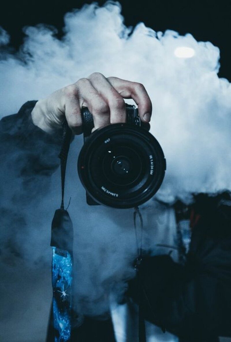 A hand holding a DSLR camera with a blue, starry camera strap, surrounded by a thick cloud of smoke in a dark environment. The focus is on the camera lens, giving a dramatic, mysterious feel to the image.