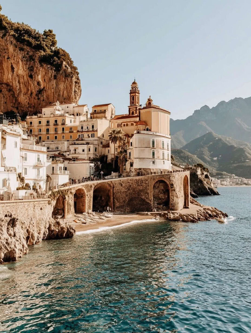 Romantic European Amalfi Coast seaside village perched on a mountainside, with elegant buildings, arched coves carved into the rocks, and sparkling water below. Tailored Oasis Travel Co.