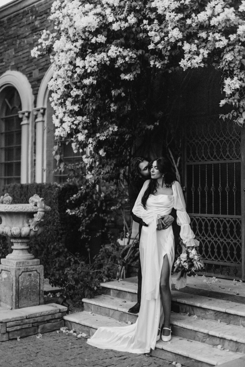 black and white of groom holding bride from behind giving neck kisses under an archway of flowers
